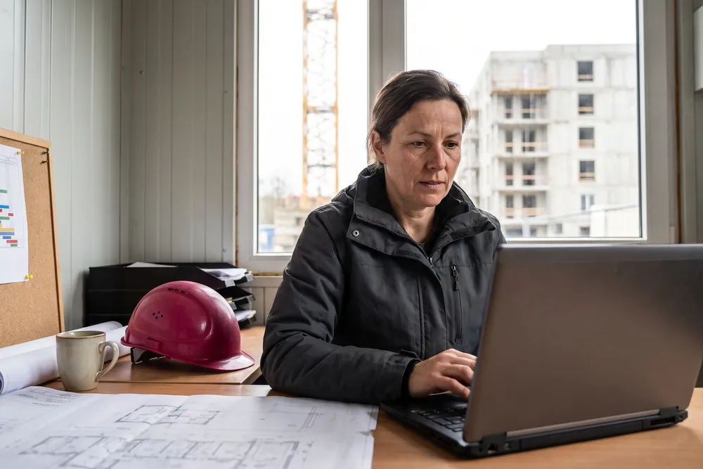 Bauleiterin im Baucontainer am Laptop, daneben Pläne und ihr Helm, im Fenster der Rohbau